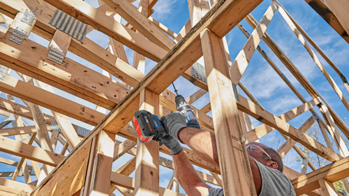 A person wearing safety glasses drills into a wooden frame with a Bosch drill, part of a wooden structure under construction, with a blue sky in the background.