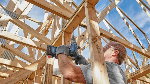 A person wearing safety glasses drills into wooden framing with a Bosch cordless drill. The drill has “CORE 18V” and “KICKBACK CONTROL” printed on it. The sky is visible in the background.