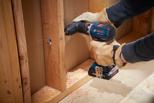 Bosch GSR18V-535FC drill being used to drive a screw into wooden studs. The drill is held by a person wearing leather work gloves.