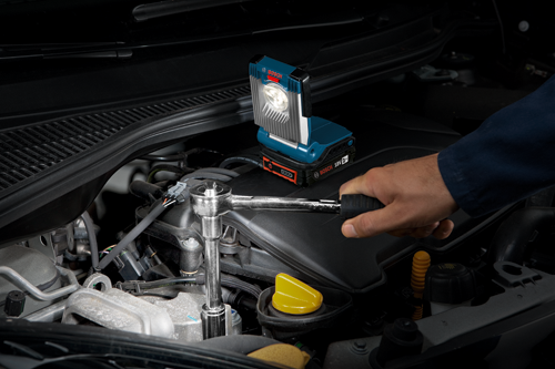 Bosch 18V 2Ah work light illuminates a person's hand using a wrench on a car engine.