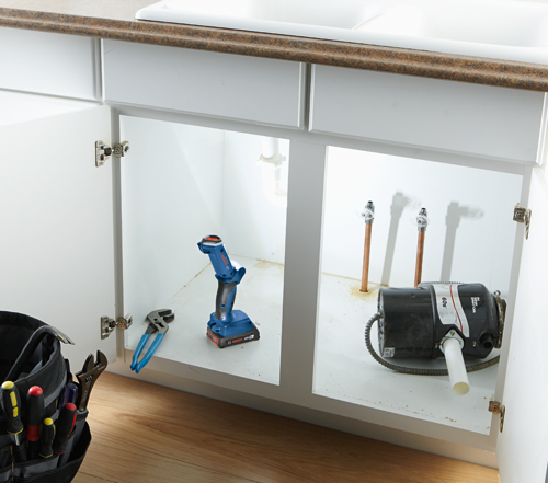 Bosch 18V 2.0Ah cordless drill, plumbing tools and a sink garbage disposal unit inside a white kitchen cabinet.
