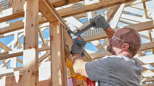 A person wearing safety glasses and gloves uses a Bosch cordless drill to work on a wood frame construction.