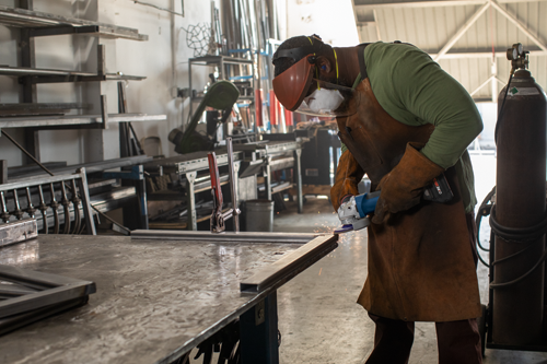 A Black person wearing protective gear, including a mask, face shield, gloves, and apron, uses a grinder on a metal bar.