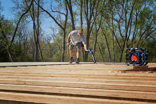 A person wearing sunglasses and work clothes uses a caulking gun on a wooden surface, with trees in the background, a portable light and other tools visible.