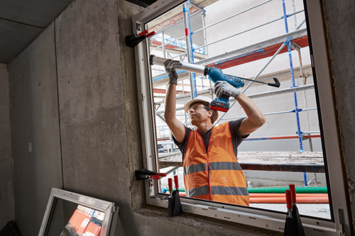 A construction worker in an orange vest, safety glasses, and hard hat using a Bosch cordless caulking gun, CORE18V battery.