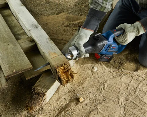 Bosch reciprocating saw cutting wood. A person wears gloves and jeans. The saw has "Bosch" and "18V" printed on it. The wood is part of a wooden structure, and the person is kneeling on the sand.