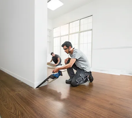 Man kneeling on a wood floor uses a Bosch CORE18V cordless reciprocating saw to cut a baseboard, with another person in the background.