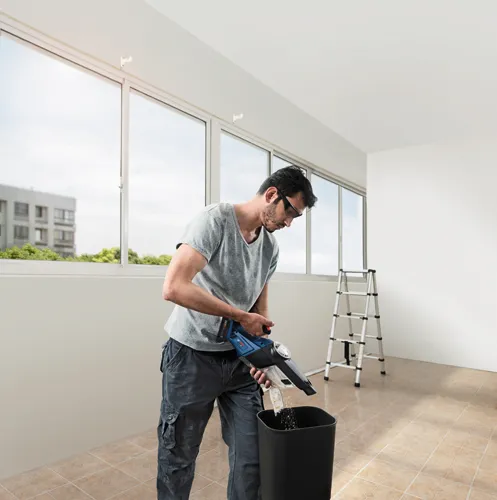 A person wearing glasses empties a Bosch vacuum cleaner into a black trash can.