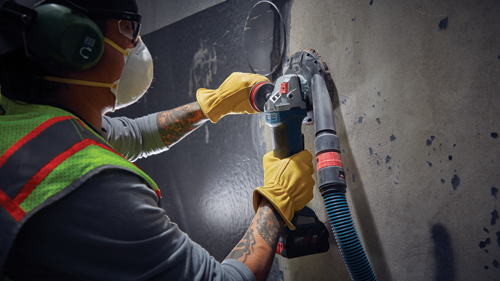 Person wearing protective gear using a Bosch power tool to sand a wall. The tool is connected to a vacuum hose.