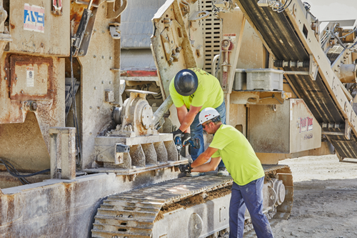 Two people wearing hard hats and safety shirts working on heavy machinery, using a power drill. The machinery has the "EasyTrak" logo with "FT4250" visible.