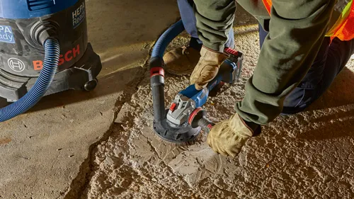 Bosch HEPA Ready vacuum and angle grinder used by a person wearing gloves to grind concrete, with hose attached.