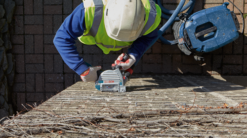 Person wearing a reflective vest and gloves uses a Bosch power saw on a brick surface, with a blue vacuum cleaner nearby.