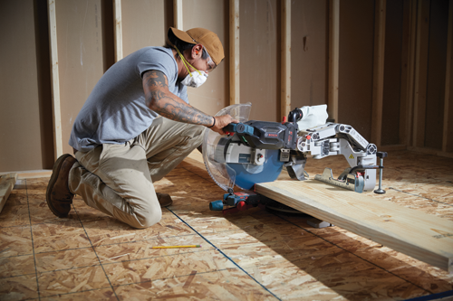 A person with tattoos kneels, using a Bosch miter saw to cut a wooden plank.