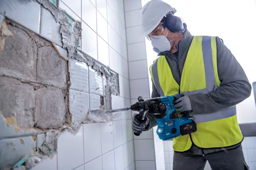 A person wearing safety gear, including a hard hat, safety glasses, and a respirator, uses a Bosch hammer drill to break apart a tiled wall.