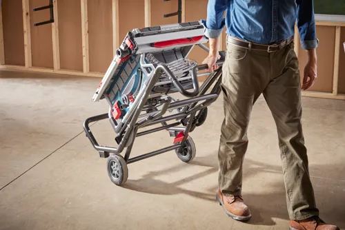 Man in jeans shirt and work pants transporting a Bosch table saw on wheels.