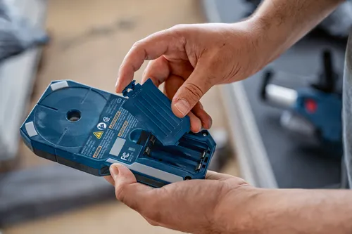 Bosch GMS120-27, with the battery compartment open, revealing the battery slots and the cover with "OPEN" text. The device has warning text, a circular feature, and is held by a person.