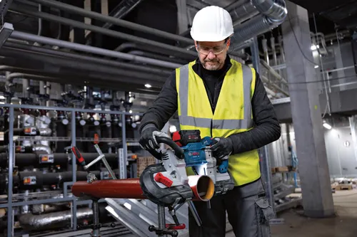 Man wearing white hard hat and safety glasses, cutting a pipe with a Bosch GCB18V-6 cordless band saw.
