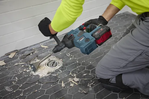 Bosch 18V Hammer Drill, a person kneels with a Bosch tool to remove tiles from a floor. The drill is powered by an "AMP Share" 18V 6.0 Ah battery.