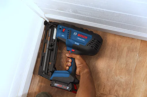 Bosch GNH18V-16MA 16 GA Finish Nailer held by a person with dark skin, in front of a white frame on a wooden floor.