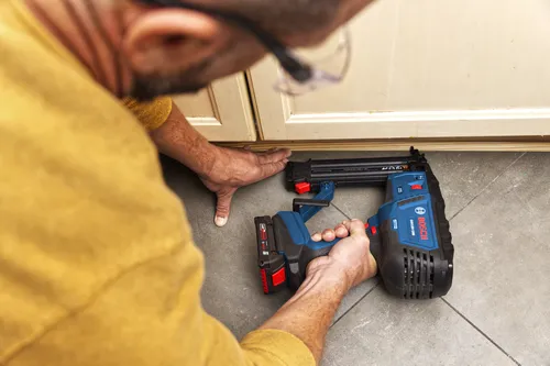 A person wearing a yellow shirt and safety glasses uses a Bosch GNH18V-18M nailer, a 18-GA tool.
