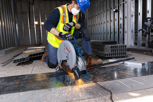 Person wearing a safety helmet, mask, and vest using a Bosch cut-off saw to cut a metal bar, with sparks flying.