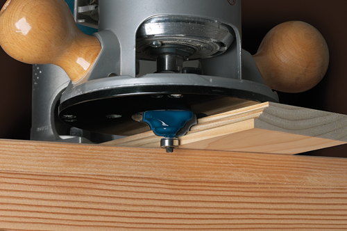 A wood router with a blue bit is cutting a decorative edge on a wooden board, with wooden handles visible.