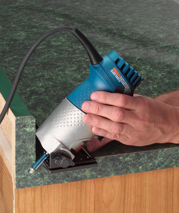 A person's hands hold a blue and silver electric tool against a green surface; the tool is cutting a corner of the surface.