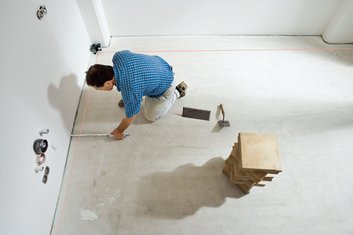 A person wearing a blue plaid shirt and tan pants kneels on a concrete floor, measuring a wall with a ruler near a red laser line.