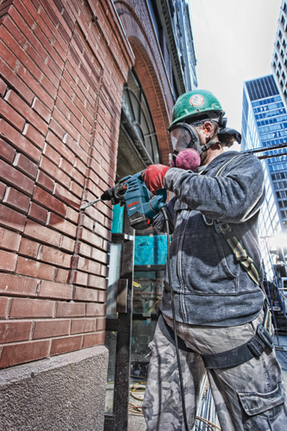 Una persona que lleva un casco verde y un respirador utiliza un Boschhammer RH328VC de Bosch para perforar una pared de ladrillo. La persona lleva una chaqueta gris, pantalones de camuflaje y guantes rojos.