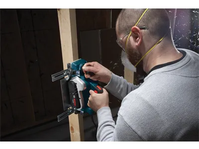 Persona que lleva gafas de seguridad y un respirador utiliza una herramienta eléctrica para cortar madera. La herramienta tiene "Bosch" impreso en el lateral.