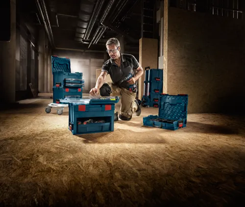 Hombre con gafas de seguridad y ropa de trabajo sosteniendo un taladro eléctrico mientras trabaja con varios organizadores de cajas de herramientas Bosch azules y rojas.