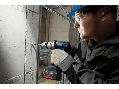 Persona que lleva gafas de seguridad, orejeras y un casco de seguridad utilizando un taladro Bosch para perforar un agujero en una pared de hormigón.