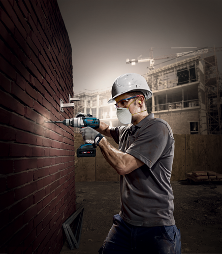 Una persona que lleva gafas de seguridad, una mascarilla antipolvo y tapones para los oídos taladra una pared de ladrillo con un taladro inalámbrico etiquetado como 'Bosch LITHIUM-ION' y 'CORE18V'.
