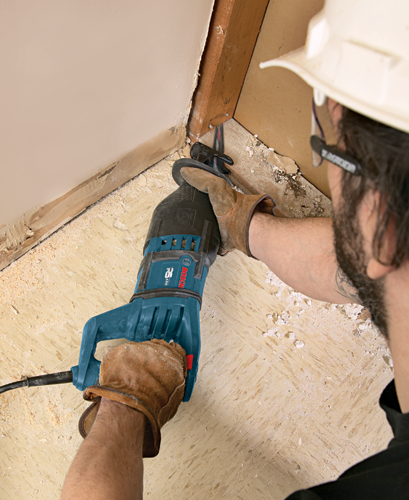 Una persona que lleva guantes de trabajo, gafas de seguridad y un casco blanco utiliza una sierra recíproca para cortar una estructura de madera. La sierra es un modelo Bosch.