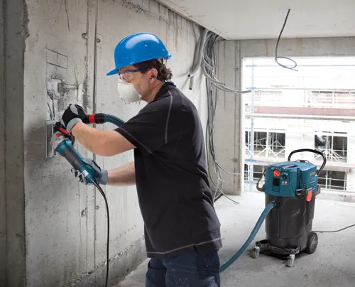 Una persona que lleva equipo de protección utiliza una amoladora Bosch en una pared de hormigón, con un aspirador Bosch cerca conectado con una manguera.