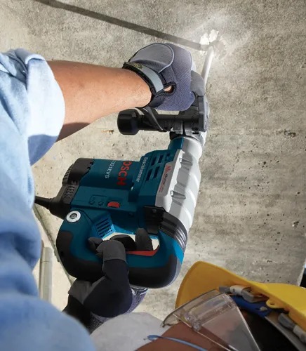 Bosch 11321EVS Boschhammer, sostenido por una persona que lleva guantes de trabajo, taladrando hormigón. La persona lleva una camisa azul y gafas de seguridad. Un casco amarillo es parcialmente visible.