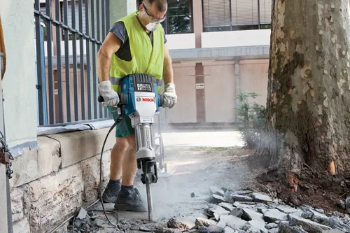 Persona operando un martillo perforador Bosch "BRUTE TURBO", con gafas de seguridad, guantes y un chaleco de alta visibilidad.