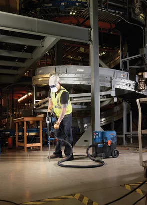 Una persona que lleva un casco, una mascarilla y un chaleco de alta visibilidad opera un martillo perforador Bosch conectado a una aspiradora con el texto "HEPA Ready", en interiores.
