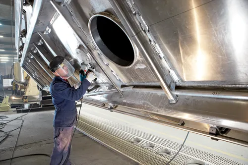 Persona con equipo de protección utilizando una herramienta eléctrica en una estructura metálica grande, con el texto visible "QSV" y "M 179040 6018".