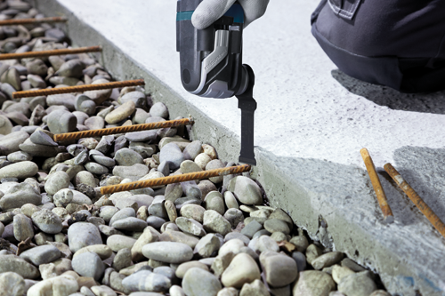 Una mano enguantada utilizando una multiherramienta StarlockMax para cortar hormigón cerca de varillas de refuerzo y guijarros.