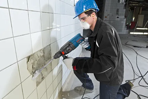 Persona utilizando un martillo rotativo Bosch BULLDOG Xtreme para retirar azulejos de la pared, con gafas de seguridad, mascarilla antipolvo, tapones para los oídos, guantes y casco azul.
