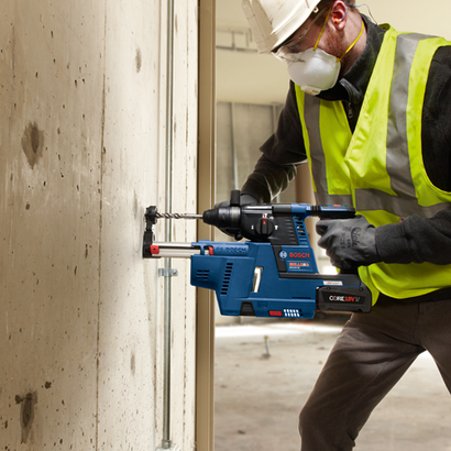 Aquí está la traducción:

Bosch BULLDOG GBH18V-26 martillo rotativo inalámbrico siendo utilizado por una persona que lleva gafas de seguridad, un respirador, un casco y un chaleco de alta visibilidad.