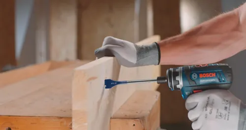 Hombre usando taladro Bosch GSR12V-140FC para perforar un agujero en una viga de madera; texto en el taladro: “BOSCH”.