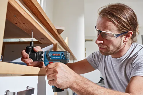 Hombre con gafas de seguridad utilizando un taladro Bosch GSR12V-140FC para ensamblar escaleras de madera.