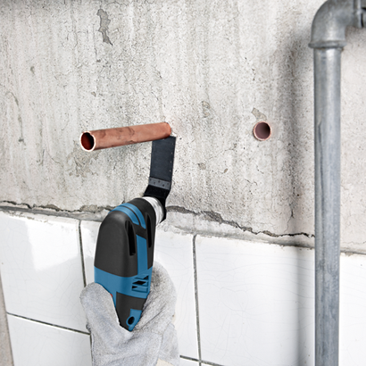 Herramienta multifunción oscilante Bosch cortando tubería de cobre en una pared. Una mano con un guante de trabajo sujeta la herramienta.