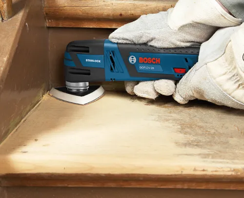Bosch GOP12V-28 STARLOCK multiherramienta, con una lija de grano, sostenida por manos enguantadas.
