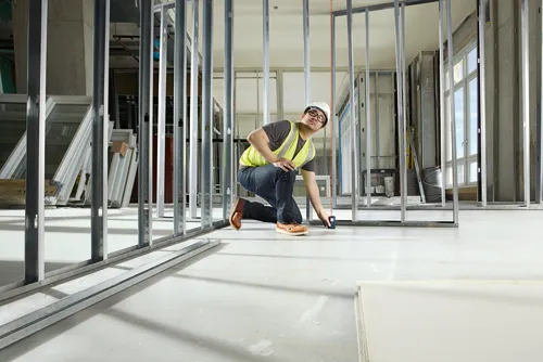 Persona asiática que lleva gafas de seguridad, un casco blanco y un chaleco amarillo, utiliza un medidor de distancia láser en una obra de construcción.