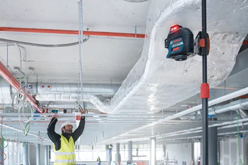 Una persona que lleva un chaleco de seguridad amarillo y un casco de seguridad está trabajando con un nivel láser Bosch Professional GLL 3-80 montado en un poste en una obra.
