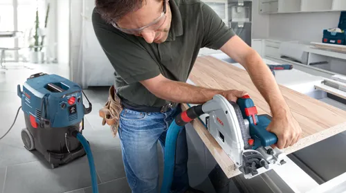 Persona que lleva gafas de seguridad utiliza una sierra circular, conectada a un aspirador Bosch para cortar una pieza de madera.