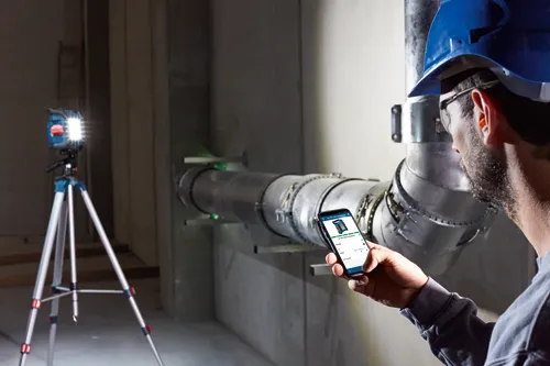 Hombre con casco de seguridad sosteniendo un teléfono con la aplicación GLI 18V-1200 C Professional, un nivel láser montado en trípode y una tubería metálica.